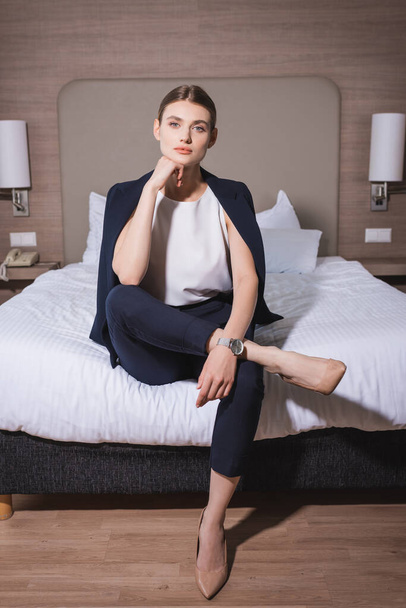 businesswoman in suit looking at camera and sitting on bed in hotel  - Фото, зображення