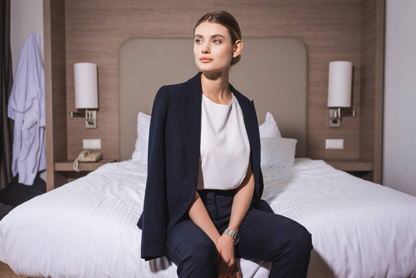 jonge zakenvrouw in pak kijken weg en zitten in bed op hotel  - Foto, afbeelding