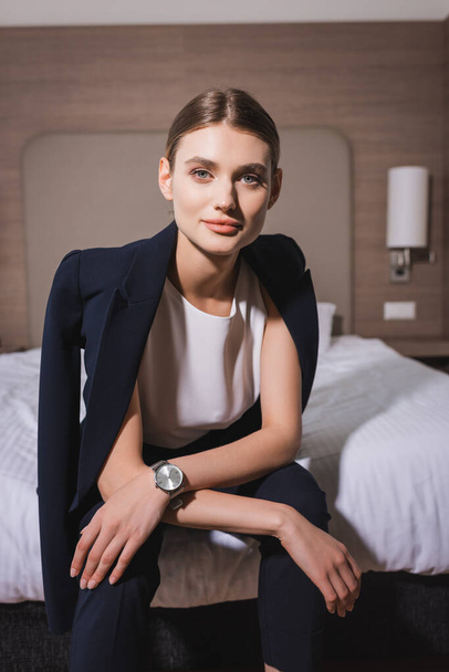 young businesswoman in formal wear looking at camera and sitting on bed in hotel room - 写真・画像