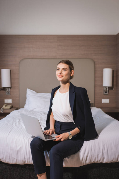 dreamy woman in suit sitting on bed with laptop in hotel room  - Foto, Bild