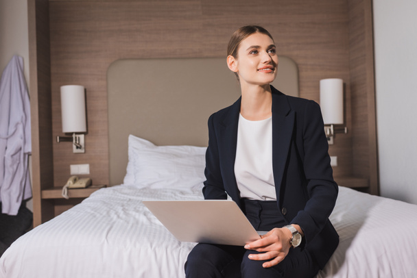 радостная женщина в костюме сидит на кровати с ноутбуком и смотрит вдаль в номере отеля  - Фото, изображение