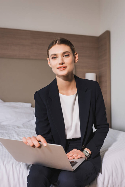 joyful woman in suit sitting on bed with laptop and looking at camera in hotel room  - Foto, Bild