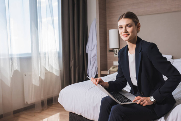 pleased young woman in suit sitting on bed with laptop in hotel room  - Zdjęcie, obraz