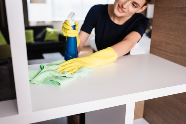 selective focus of maid in rubber gloves holding spray bottle and rag while cleaning shelf in hotel room  - Фото, зображення