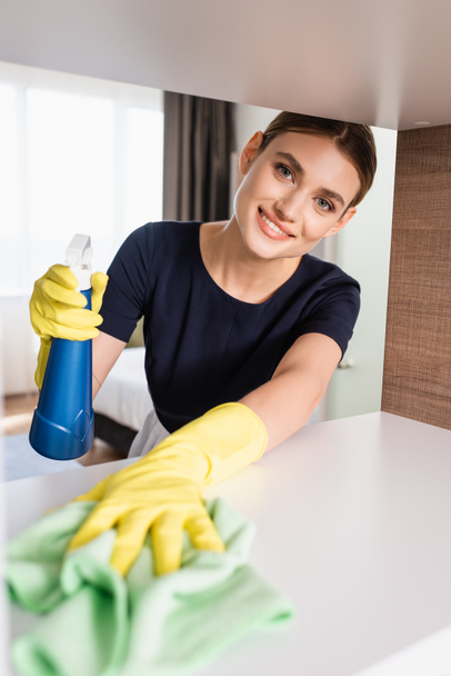 selective focus of joyful maid in rubber gloves holding spray bottle and rag while cleaning shelf in hotel room  - Фото, зображення