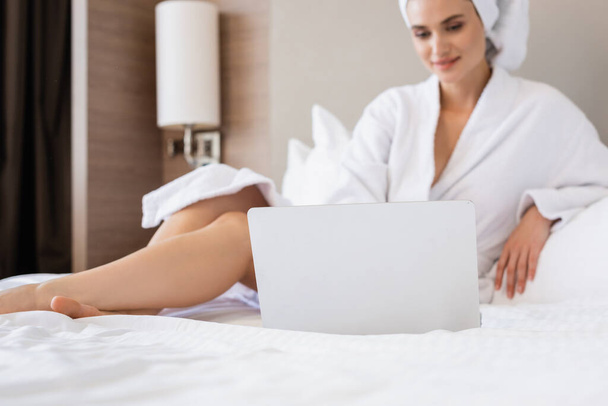 Selektiver Fokus der jungen Frau im Bademantel mit Laptop auf dem Bett - Foto, Bild