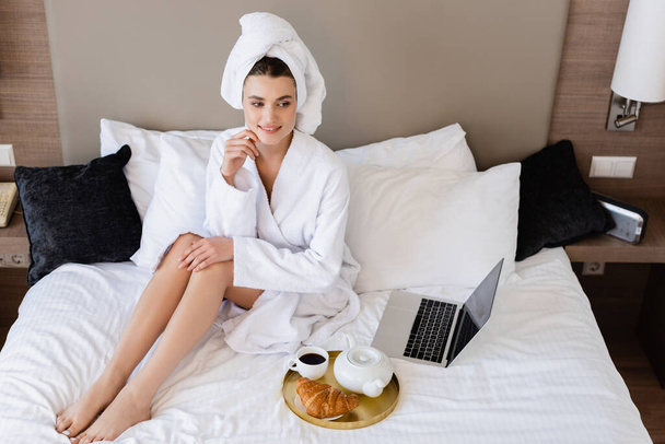 high angle view of young woman in bathrobe sitting near laptop and breakfast tray on bed - Foto, imagen