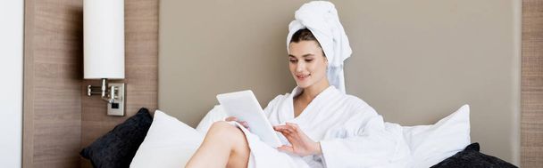 panoramisch beeld van de vrouw in handdoek en witte badjas met behulp van digitale tablet in de hotelkamer - Foto, afbeelding
