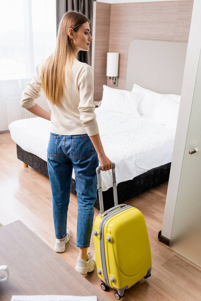 νεαρή γυναίκα με τζιν στέκεται με κίτρινες αποσκευές στο δωμάτιο του ξενοδοχείου - Φωτογραφία, εικόνα