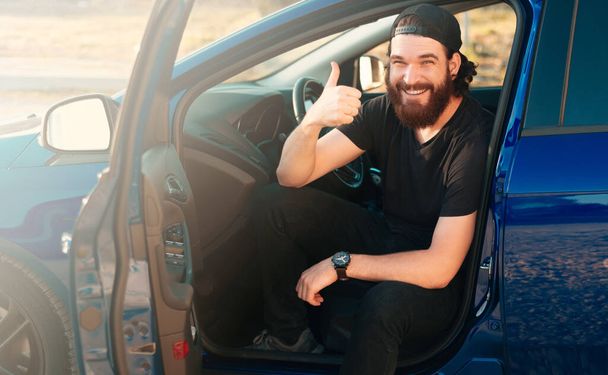 Fotografie veselého vousatého muže sedícího ve svém novém autě a ukazujícího palce nahoru - Fotografie, Obrázek