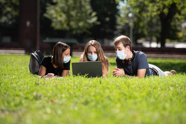 Powrót do unibersity podczas Coronavirus lub Covid-19. Trzech studentów z laptopem leżącym i rozmawiającym w parku w masce na twarz. - Zdjęcie, obraz