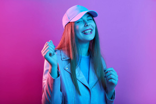 Fashionable woman stands smiling against pink neon wall, having fun in stylish outfit, beautiful girl with long hair. Cheerful young model looking directly at camera. - Zdjęcie, obraz