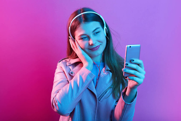 Adorable attractive teenage girl listening music in headphones isolated over pink neon background, touches her ear, looking at smart phone screen, wearing leather jacket. - Fotografie, Obrázek