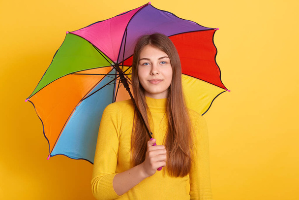 Attractive young woman standing under multicolored umbrella and looking directly at camera, wearing yellow jumper, girl with long hair has calm facial expression. - Foto, Imagen