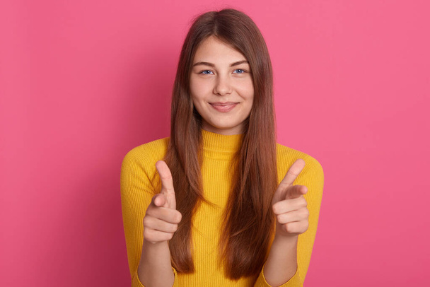Smiling pretty young woman pointing to camera with flirting facial expression, looks happy, expressing positive emotions, I choose you, wearing yellow jumper against pink wall. - Фото, зображення