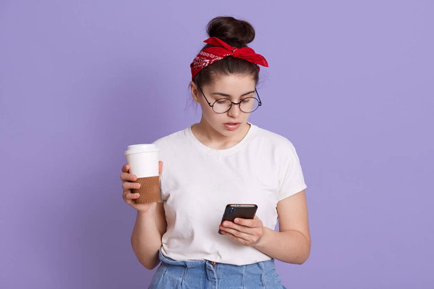Attractive brunette young caucasian woman holds modern mobile phone and coffee to go, sends text messages in online chat, wears white casual t shirt and red hairband, poses over lilac background. - Foto, Imagem