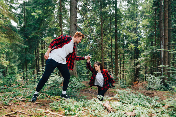 若い男は女性がハイキングに登るのを助ける。赤いシャツを着たカップルが森を歩き、男は立ち上がりで女性の手をサポートします。ハイキング中の美しいカップル - 写真・画像