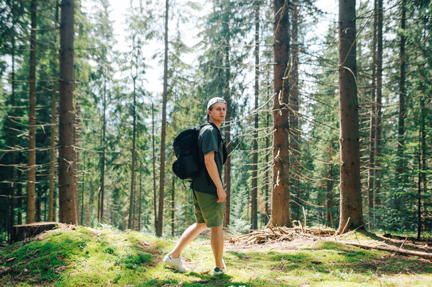 Jóképű fiatal férfi túrázó egy túrán a hegyekben áll egy érintetlen tűlevelű erdőben, és néz a kamera egy komoly arcot.Turista utazik keresztül az érintetlen gyönyörű erdőben, és pózol - Fotó, kép