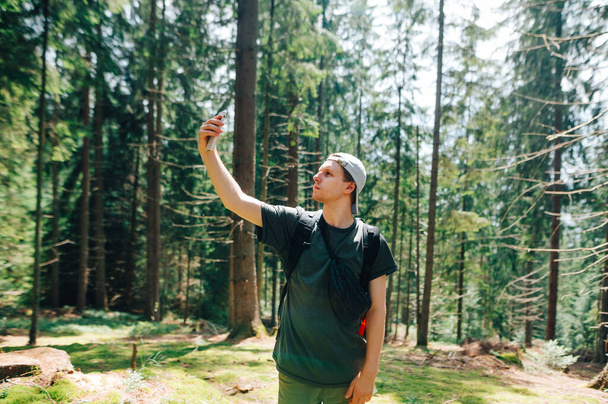 Jonge mannelijke toerist op zoek naar een mobiel netwerk op een smartphone in een prachtig bos, tilde een telefoon op en kijkt met een serieus gezicht naar het scherm. Guy gebruikt het internet op een smartphone op een wandeling - Foto, afbeelding
