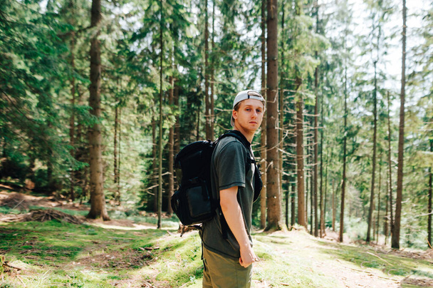 Handsome young man in casual clothes and with a backpack stands in a sunny forest and land with moss and poses for the camera in the camera with a serious face. Guy tourist in a forest hike - Photo, Image
