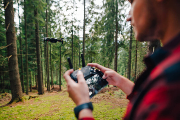 Ο άνθρωπος ελέγχει ένα τηλεκατευθυνόμενο από τηλεχειριστήριο στο δάσος, εστιάζοντας σε ένα τηλεκατευθυνόμενο εν πτήσει. Ιστορικό. Ένας τουρίστας εκτοξεύει ένα drone για να φωτογραφίσει το δάσος. - Φωτογραφία, εικόνα