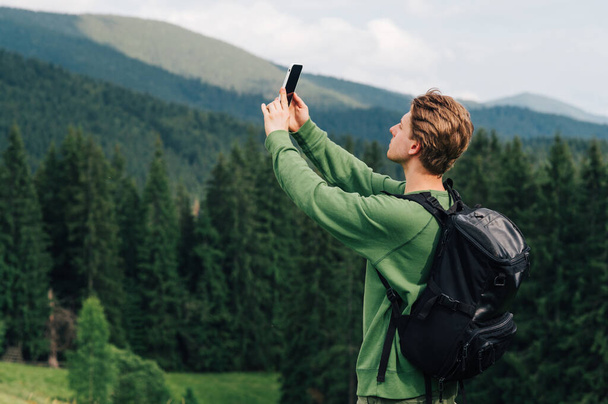 Günlük kıyafetli genç bir adam cep telefonu şebekesini elleri havada bir şekilde yakalıyor. Turist, dağ ormanlarının ve dağların arka planında yürüyüş yaparken selfie çeker.. - Fotoğraf, Görsel