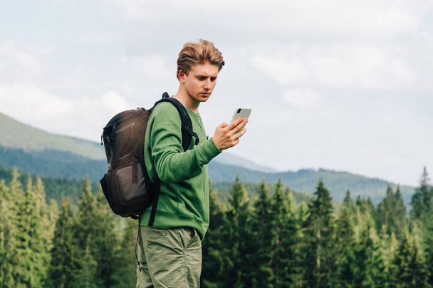 Skoncentrowany młody człowiek z plecakiem i w luźnych ubraniach używa smartfona na tle pięknego krajobrazu z górskim lasem iglastym. Facet używa Internetu w górach. - Zdjęcie, obraz