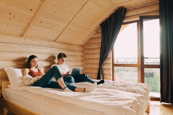 Young couple lying on a bed in an apartment with a wooden interior, a woman uses a smartphone and a man works on a laptop. Leisure in a hotel room. - Фото, зображення