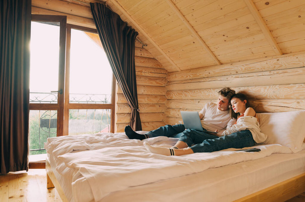 Young beautiful couple watching a movie on a laptop while lying on a bed in a wooden apartment. Man and young woman using laptop on vacation, relaxing in hotel - Foto, imagen