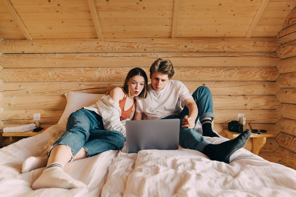 Překvapený mladý pár ležící v posteli v bytě s dřevěným interiérem a dívající se na obrazovku notebooku s šokovanými tvářemi. Šokovaný pár sledující film v posteli - Fotografie, Obrázek