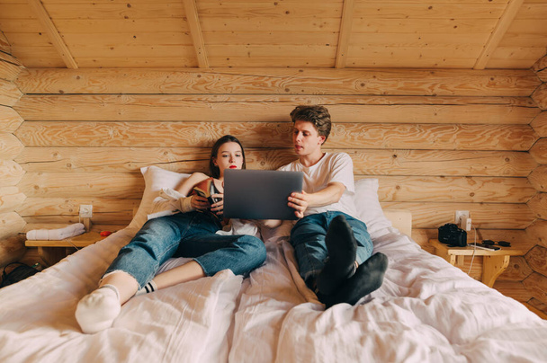 Young beautiful couple lying on bed in wooden apartment and resting, young man showing laptop to woman with book. Leisure at the hotel. - Φωτογραφία, εικόνα