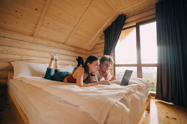 Happy young couple lying on bed in hotel room and using internet on laptop with smiles on their faces. Woman and man watching movie on laptop in wooden apartment - Fotografie, Obrázek