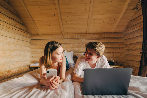 明るい若いカップルがベッドの上に横たわっている木製のインテリア、スマートフォンを持つ女性とラップトップを持つ男が顔に笑顔でお互いを見て. - 写真・画像