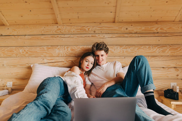 Positive young man and woman couple watching movie on laptop, lying in wooden apartment on bed. Couple relaxing watching a movie at the hotel. - Фото, изображение