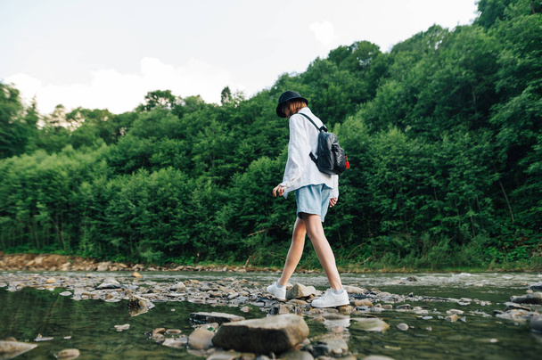 Dívka ve stylovém neformálním oblečení se prochází po skalách na řece v horách. Žena turista procházky po krbu na řece lesní - Fotografie, Obrázek