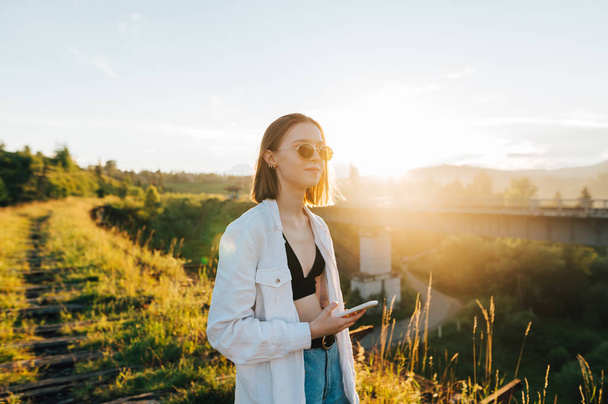 Pretty girl in sunglasses and casual clothes stands on an old railway bridge with a smartphone in hand, looking to the side on a background of sunset. - Foto, afbeelding