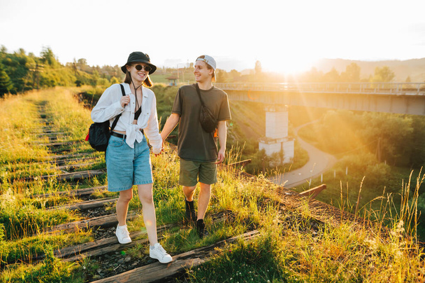 young couple walks on the old railway bridge against the backdrop of the sunrise and smiles, wearing stylish summer clothes. Young tourists go holding hands on a viaduct in the mountains - Photo, Image