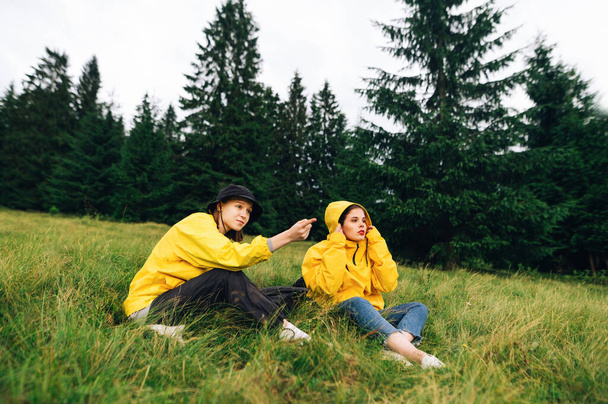Women serious tourists in yellow raincoats resting on the grass on a hike, the girl points her finger at the route. Tourists rest on a meadow in the mountains on a hike - Photo, Image
