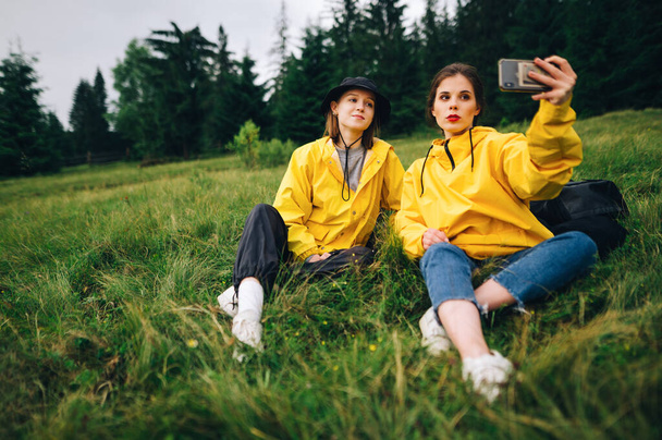 Beautiful girl in a yellow raincoat taking a selfie on a mountain hike sitting on a meadow on a background of forest. Tourists take a selfie at a halt during the hike - Foto, immagini