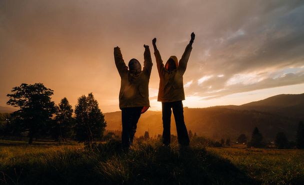 Two hikers in yellow raincoats stand on a meadow on a background of sunset in the mountains with raised arms. Silhouette of joyful tourists on a background of sunset in the rain - Foto, Imagem