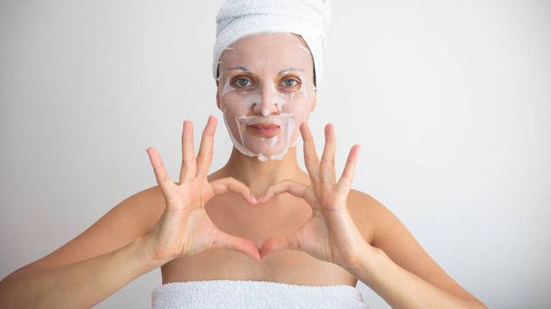 Женщина завернутая в полотенце с белой косметической тканью маска для лица дает символ сердца руками.  - Фото, изображение