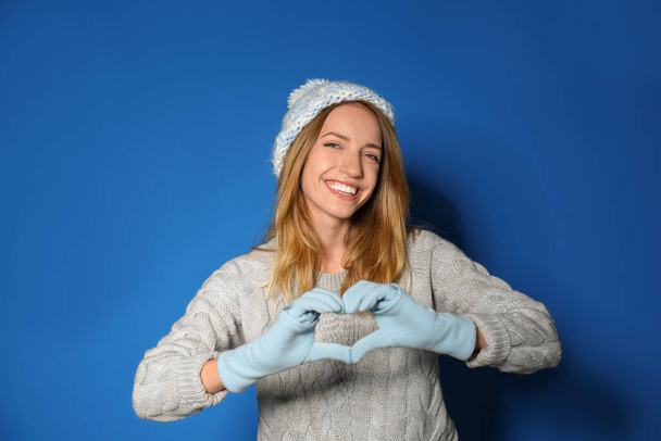 Happy young woman in warm sweater making heart with hands on blue background - Photo, Image
