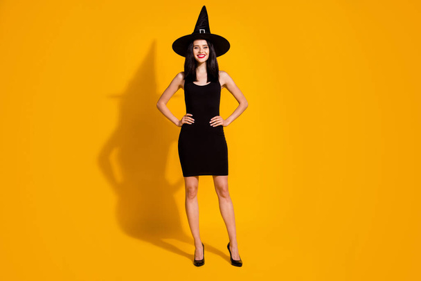 Full length photo of charming pretty young witch lady hands hips self-assured beaming smiling funny event wear black headwear high heels dress isolated bright yellow color background - Fotografie, Obrázek