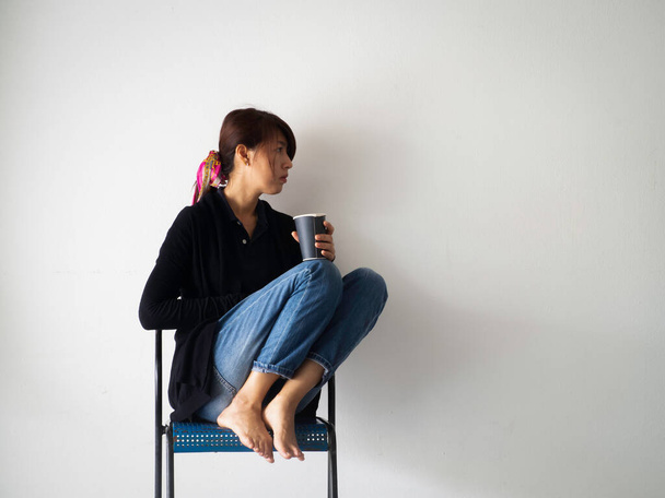 Vrouw zit op stoel tegen de muur, houdt koffiebeker in de hand met ongelukkig gevoel, beproefd en verdrietig gevoel - Foto, afbeelding