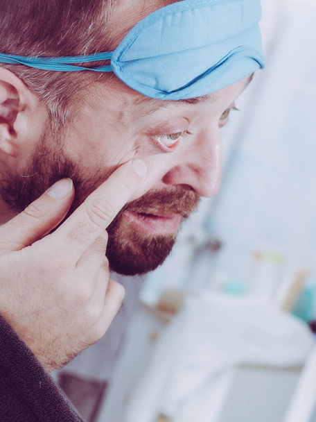 Funny adult man wearing eyemask on forhead having troubles with waking up. Standing in front of mirror investigating imperfections on his face. - Fotografie, Obrázek