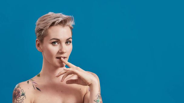 Retrato de uma jovem atraente mulher tatuada semi-nua com pele perfeita olhando brincalhão, segurando o dedo na boca isolado sobre fundo azul - Foto, Imagem