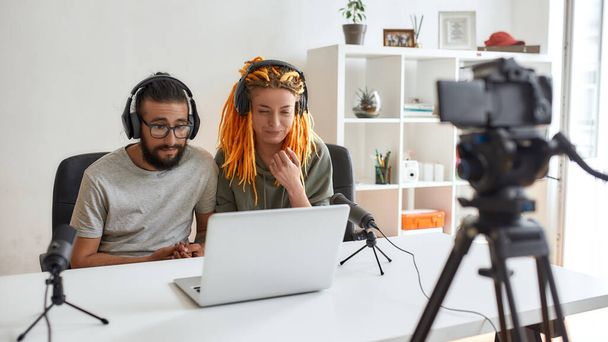 Couple of bloggers, young man and woman in headphones watching something on laptop, making faces while recording reaction video blog or vlog - Φωτογραφία, εικόνα