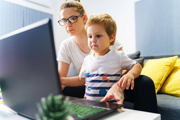 Mały chłopiec stojący przy matce przed laptopem w domu - Mały chłopiec korzystający z komputera do oglądania wideo online lub wykonywania połączeń - Zdjęcie, obraz