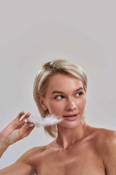 Portrait of beautiful young female blonde with perfect fresh skin looking aside, touching her face with white feather while posing isolated over grey background - 写真・画像