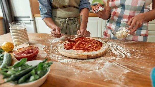 Cropped shot of couple making pizza together. Man in apron adding, applying tomato sauce on the dough while woman helping him in the kitchen. Hobby, lifestyle - Photo, Image